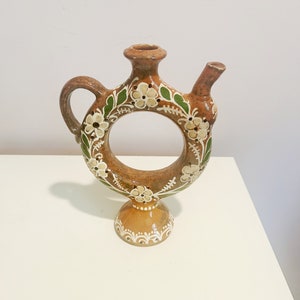 Puede incluir: Una jarra de cerámica marrón con un asa en forma de anillo y un diseño floral. La jarra tiene un pequeño pico y una base con un patrón decorativo.