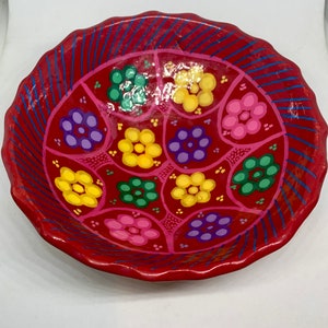 Pode incluir: Uma tigela de cerâmica vermelha com uma borda ondulada e um design decorativo de flores coloridas. As flores são pintadas em tons de amarelo, verde, roxo e rosa.