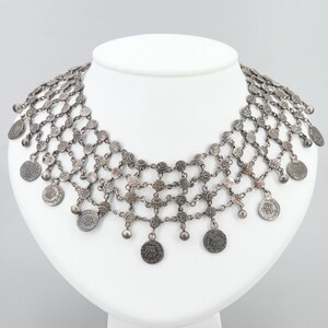 May include: A silver necklace with a delicate chain and multiple rows of coin-shaped pendants. The coins have intricate designs and are connected by small, round beads.