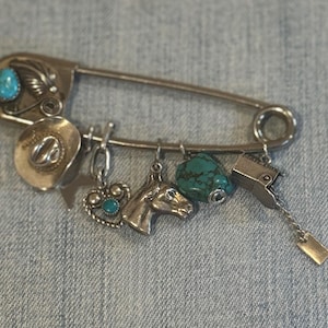 Puede incluir: Un imperdible plateado con dijes de temática occidental y detalles turquesa. Los dijes incluyen un sombrero de vaquero, un corazón, una cabeza de caballo, una cuenta turquesa y un buzón. El imperdible tiene una piedra turquesa.
