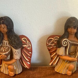 May include: Two ceramic angel figurines with dark hair, holding musical instruments. The angels have tan skin, wearing cream and yellow dresses, and red and cream wings. One holds a drum, the other a harp.