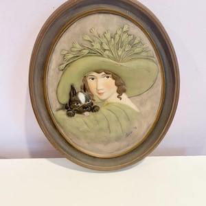 May include: A framed oval-shaped wall plaque with a 3D relief image of a woman wearing a large green hat with a dog. The woman is looking at the viewer and the dog is looking at the woman. The plaque is signed "Susan '78".