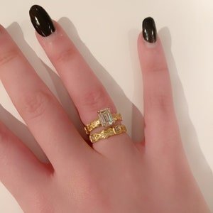 May include: A close-up of a hand wearing two gold rings. The top ring features a large, clear emerald-cut gemstone. The bottom ring has a textured band with smaller gemstones.