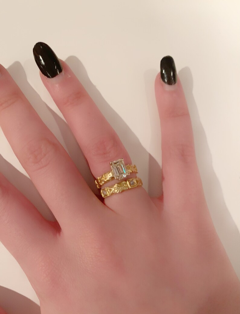May include: A close-up of a hand wearing two gold rings. The top ring features a large, clear emerald-cut gemstone. The bottom ring has a textured band with smaller gemstones.