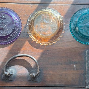 May include: Three antique glass plates in purple, amber, and teal, each with a detailed embossed design. The purple plate features a frog, the amber plate a rooster, and the teal plate a bird. A metal handle and hinge are also visible.