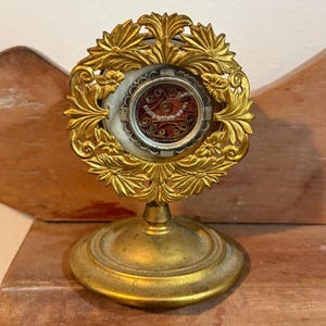 Pode incluir: Um relicário dourado com um design floral ornamentado. O relicário apresenta um centro circular com um interior vermelho e branco, montado numa base de pedestal. A peça é provavelmente vintage ou antiga.