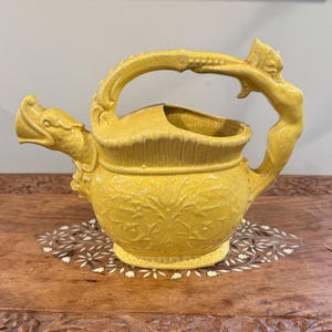 May include: A yellow ceramic pitcher with a dragon-shaped spout and a handle featuring a sculpted figure. The pitcher has a textured surface with floral patterns and a wide opening. The pitcher is sitting on a wooden surface.