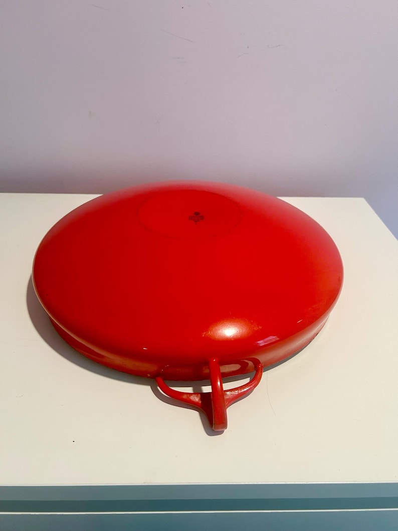May include: A red enamel cast iron pan with two handles. The pan is upside down and the bottom is visible.