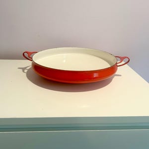 May include: A red enamel cast iron baking dish with two handles. The dish is empty and has a white interior.