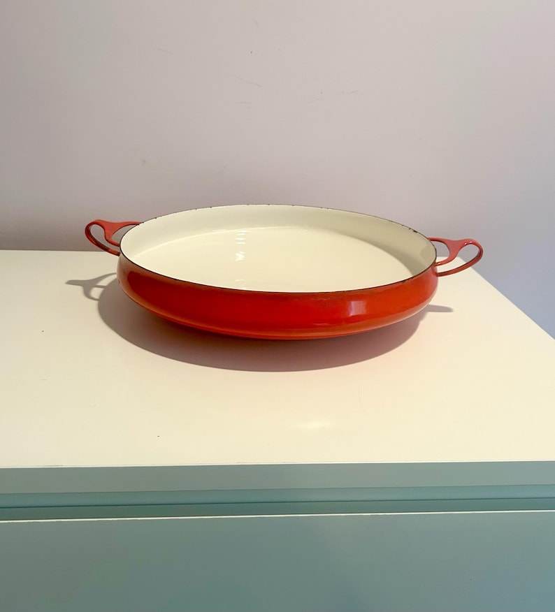 May include: A red enamel cast iron baking dish with two handles. The dish is empty and has a white interior.