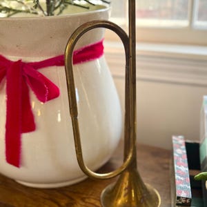 May include: A vintage brass bugle-shaped candle holder with a dried orange slice on top. The candle holder is on a wooden surface, next to a white ceramic vase with a red ribbon and a small evergreen plant.