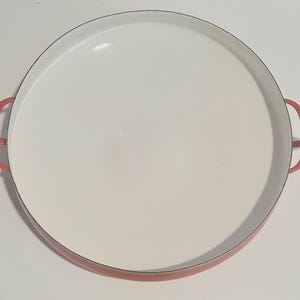 May include: Red enamel cast iron serving dish with two handles. The dish is round and has a white interior.