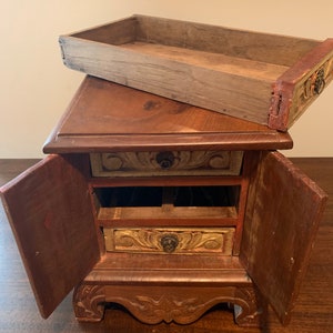 May include: A small wooden box with three drawers and two doors. The box is decorated with carved floral designs and painted in shades of brown, gold, and red.