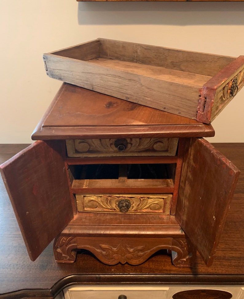 May include: A small wooden box with three drawers and two doors. The box is decorated with carved floral designs and painted in shades of brown, gold, and red.