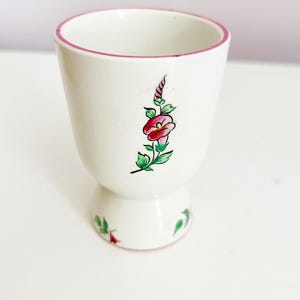May include: White ceramic egg cup with a pink rim and a floral design featuring a pink flower and green leaves.