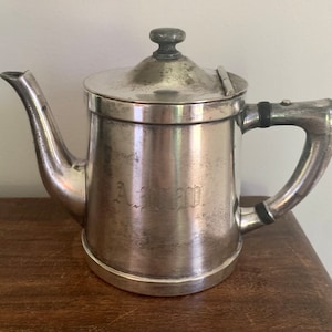 May include: A silver-colored teapot with a lid and handle. The teapot has a spout and a small knob on top of the lid. The handle is curved and attached to the side of the pot. The teapot has the engraved letters "A M" on the front.