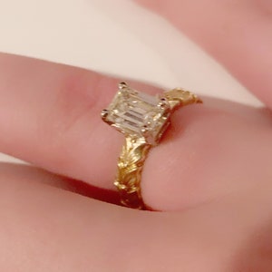 May include: A gold engagement ring with an emerald cut diamond. The band is intricately designed with a floral pattern.