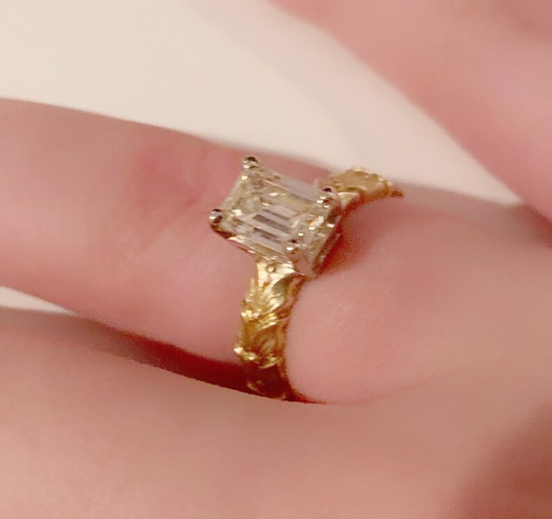 May include: A gold engagement ring with an emerald cut diamond. The band is intricately designed with a floral pattern.