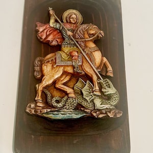 Puede incluir: Una placa de madera con una escultura en relieve de San Jorge matando a un dragón. La escultura está pintada en colores vibrantes, incluyendo rojo, amarillo, verde y azul. La placa es de color marrón oscuro y tiene un marco tallado.