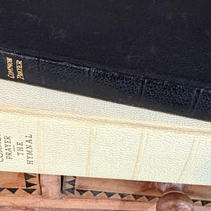 May include: Two stacked books, one black and one cream-colored, with the titles "Common Prayer" and "The Hymnal" visible. The black book has the word "Harper" on the side. The books are resting on a decorative wooden surface.