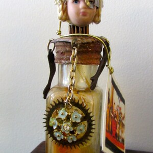 May include: A vintage glass bottle with a rusty metal lid and a doll's head on top. The doll has blonde hair, a white hat with blue flowers, and a metal eye patch. The bottle has a gold chain hanging from it and a gear with blue flowers on it. The bottle is sitting on a wooden surface. 04/27/2013 03:11