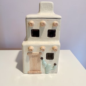 Pode incluir: Jarro de biscoitos de cerâmica branca em forma de casa de adobe do sudoeste com uma porta de madeira marrom, um cacto e um telhado verde.