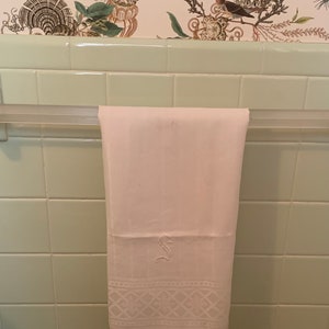 Puede incluir: Una toalla de mano blanca con una letra 'S' bordada y un borde decorativo cuelga de un toallero plateado contra una pared de azulejos verdes.