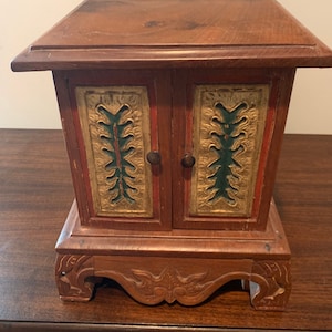 May include: A small wooden box with two doors, carved with intricate floral designs in gold, green, and red paint. The box sits on a base with carved feet.