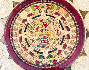 May include: A circular wooden wall hanging with a red border and a detailed design of the Aztec calendar. The calendar is made of wood and painted in various colors, including yellow, green, red, and blue.