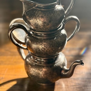 May include: Three antique silver-toned teapots stacked on top of each other. The teapots have handles and spouts. The teapots are on a wooden surface. The teapots have a vintage, tarnished appearance.