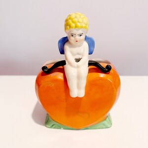 Könnte beinhalten: Ein kleiner, herzförmiger Keramik-Pflanztopf mit einer Cherubfigur darauf. Der Topf ist orange mit grünem Rand und der Cherub ist weiß mit blauen Flügeln und gelben Haaren.