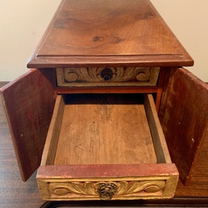 May include: A wooden box with a drawer and two doors. The box is painted in shades of red, gold, and brown. The drawer has a carved design and a brass knob. The doors are open, revealing the empty interior of the box.