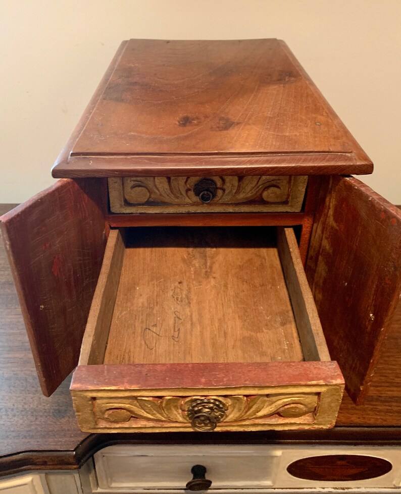 May include: A wooden box with a drawer and two doors. The box is painted in shades of red, gold, and brown. The drawer has a carved design and a brass knob. The doors are open, revealing the empty interior of the box.