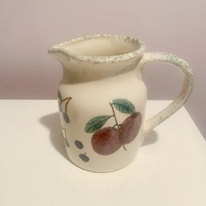 Puede incluir: Una jarra de cerámica blanca con un borde verde y un asa. La jarra tiene un diseño pintado de ciruelas y hojas.