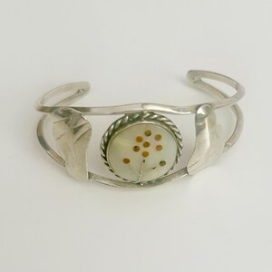 Op de afbeelding: Zilveren armband met een witte parelmoer inleg met een bruin bloemenmotief. De armband heeft een golvende vorm en twee blaadjesvormige accenten.