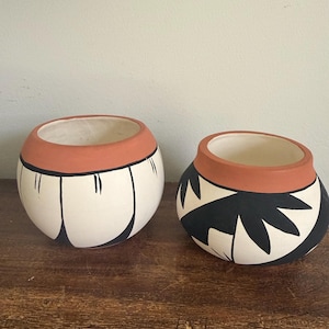 May include: Two handcrafted ceramic pots. The pots are round with a white base, a terracotta rim, and black geometric designs. One pot has vertical black lines, and the other has a black leaf-like pattern. They sit on a wooden surface.