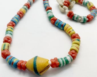 Puede incluir: Un collar de cuentas de colores con cuentas de vidrio rojas, amarillas, azules y verdes. El collar tiene una cuenta grande, amarilla y verde en el centro.