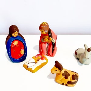 Puede incluir: Un conjunto de nacimiento con una figura con túnica azul y roja, una figura con túnica rosa, un bebé Jesús en un pesebre, un burro blanco y una vaca marrón.