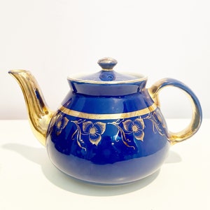Może przedstawiać: Kobaltowy niebieski czajnik z pozłacanymi zdobieniami i kwiatowymi wzorami. Czajnik ma okrągły korpus i dzióbek po lewej stronie. Uchwyt znajduje się po prawej stronie i również jest pozłacany.