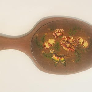 Könnte beinhalten: Handspiegel aus Holz mit Griff. Die Oberfläche des Spiegels ist mit einem Blumendesign mit orangefarbenen und gelben Blumen, grünen Blättern und einem Schmetterling verziert. Der Spiegel hat eine braune Oberfläche.