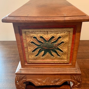 May include: A wooden box with a carved design on the front. The box is painted in shades of red, green, and gold. The box has a base with carved feet.