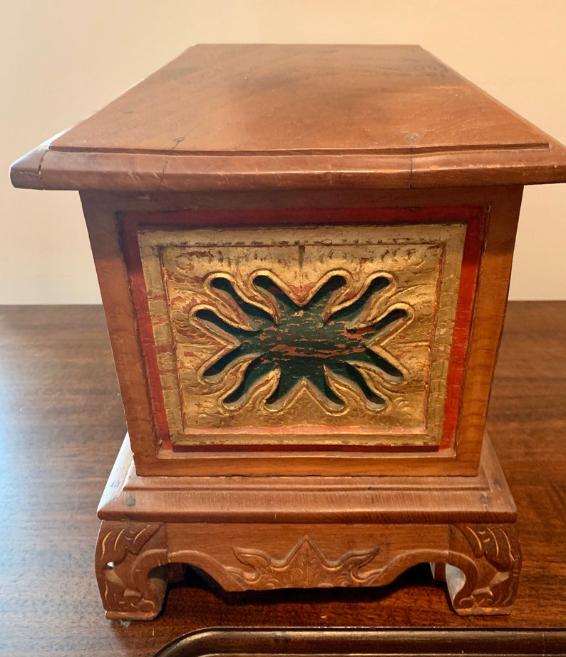 May include: A wooden box with a carved design on the front. The box is painted in shades of red, green, and gold. The box has a base with carved feet.