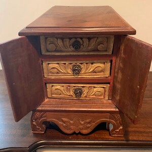 May include: A small wooden box with three drawers and two doors. The drawers and doors are carved with floral designs and have metal handles. The box is painted in shades of brown, red, and gold.