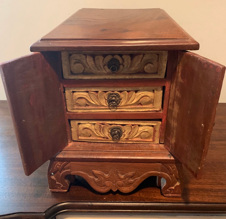 May include: A small wooden box with three drawers and two doors. The drawers and doors are carved with floral designs and have metal handles. The box is painted in shades of brown, red, and gold.