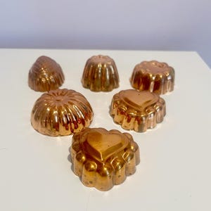Könnte beinhalten: Sechs kupferfarbene Metallformen zum Backen von Desserts. Die Formen sind in verschiedenen Formen, darunter Herz, Blume und rund.