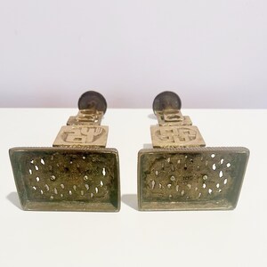 May include: A pair of gold metal candle holders with intricate designs. Each holder features a square base with a decorative pattern and a tall, slender stem with a circular top.