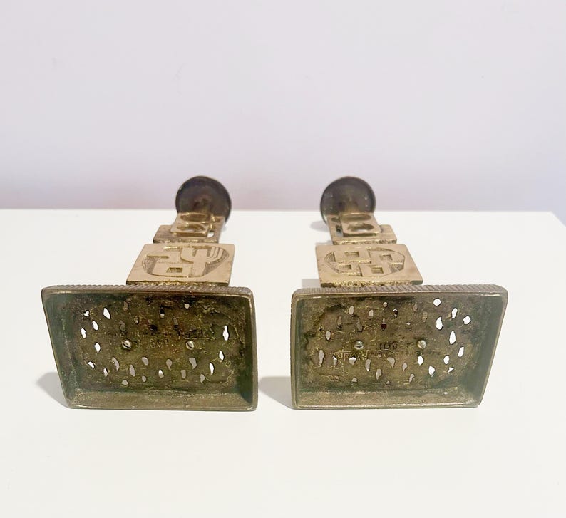 May include: A pair of gold metal candle holders with intricate designs. Each holder features a square base with a decorative pattern and a tall, slender stem with a circular top.
