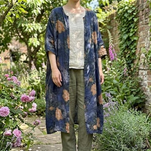 May include: A long, open-front duster coat in a dark blue fabric with a mottled texture and orange floral patterns. It is worn over a light beige top and olive green trousers, paired with brown leather ankle boots for a complete outfit.
