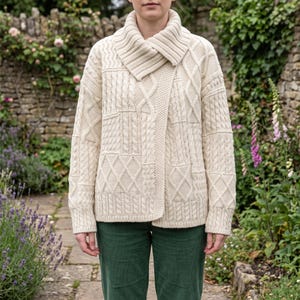 May include: An off-white, long-sleeve knit cardigan featuring a prominent shawl collar and a complex patchwork of cable knit, diamond, and vertical rib patterns. The garment has a straight hem and a thick, textured construction.