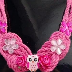 Könnte beinhalten: Eine rosa Seilkette mit einem Eulenanhänger und floralen Verzierungen. Die Kette besteht aus rosa Seil, verziert mit rosa und klaren blütenförmigen Perlen und kleinen rosa Rosen. Der Eulenanhänger ist rosa und weiß.